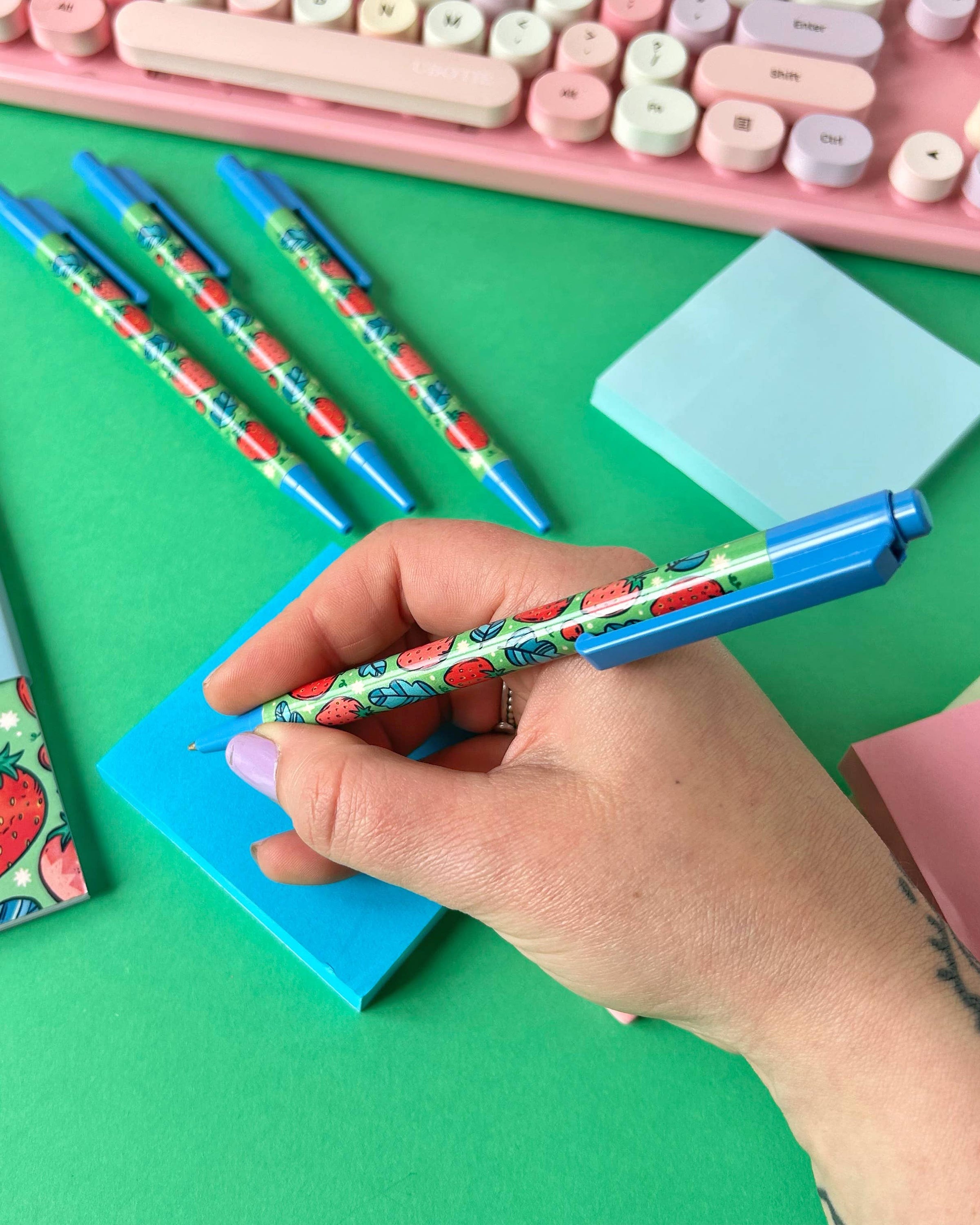 A hand holding a Rebel Romance strawberry ballpoint pen with blue accents, writing on a bright blue sticky note. A pastel pink keyboard and other pens are visible in the background. The pen features a cute all-over strawberry design by Stacey McEvoy Caunt, ideal for journaling and note-taking.