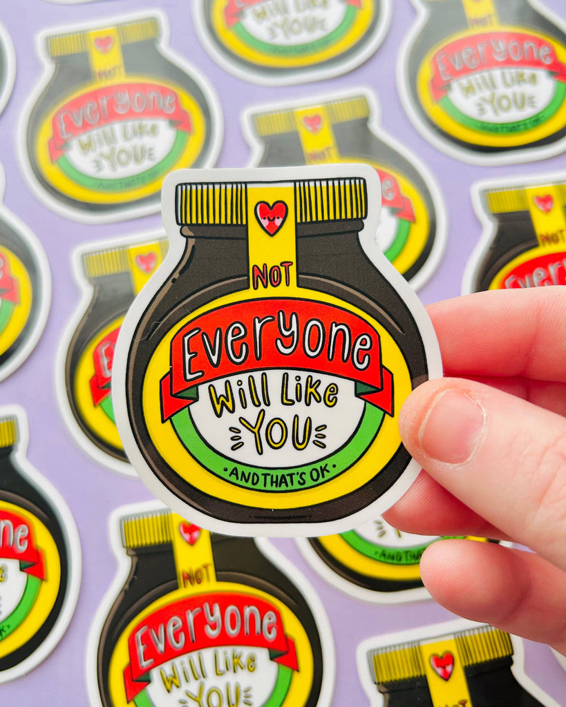 A close-up of a hand holding a single Marmite-style vinyl sticker that reads: “Not Everyone Will Like You – And That’s OK.” The sticker is surrounded by identical designs on a purple background. From Rebel Romance, a small UK brand for quirky and empowering gifts.