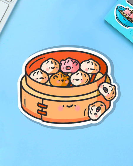 Toastedink's Steamed Buns Vinyl Sticker on a pastel blue backdrop. Features cute dumplings, tiger and pig characters nestled in a smiling bamboo steamer. Available at Rebel Romance – a UK small business celebrating quirky food art and alternative style.