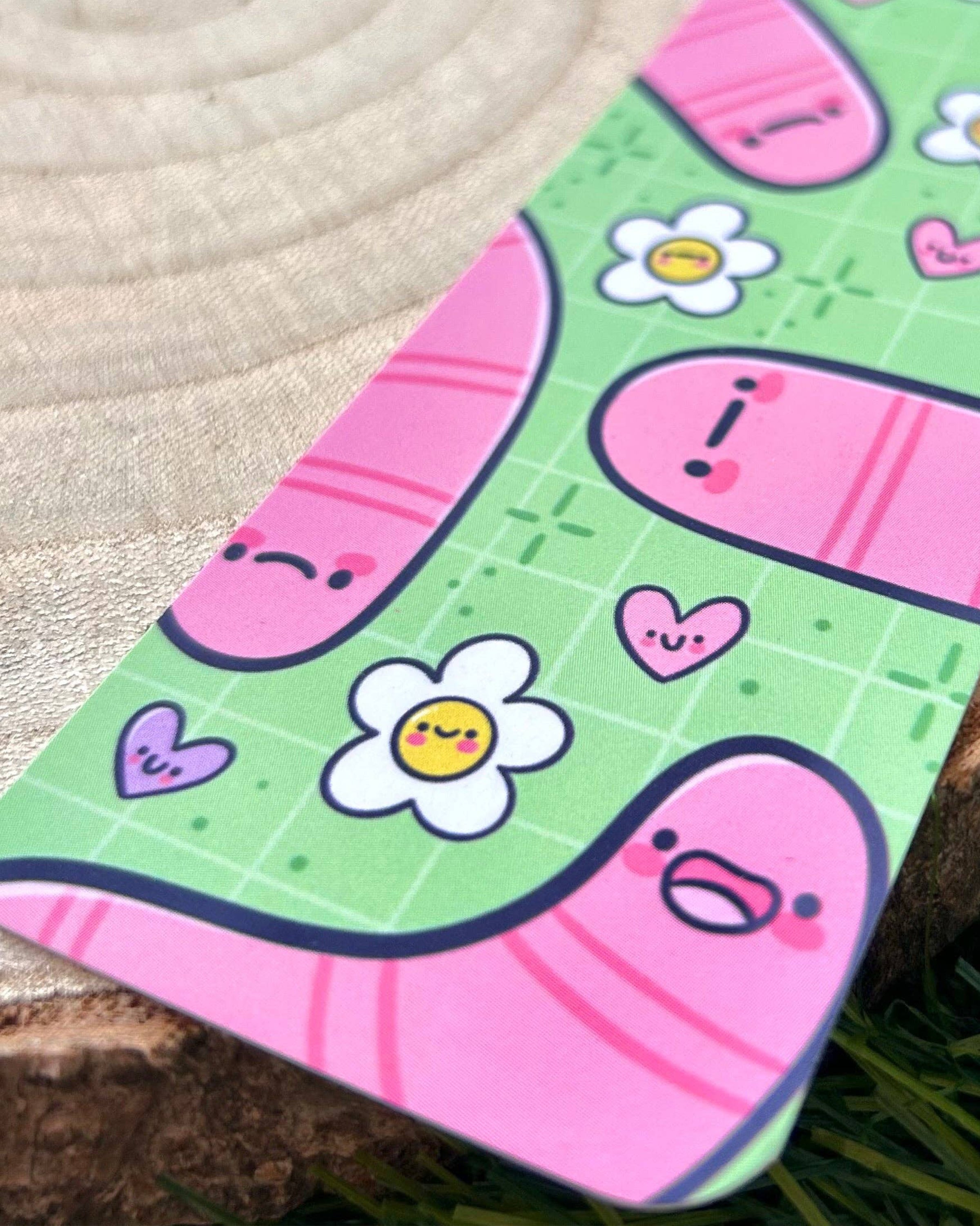 Close‑up of the Wormies Bookmark by Kelly Lou, sold by Rebel Romance. The illustrated design features pink cartoon worms with expressive faces, white daisies with smiley centres and tiny hearts on a green grid background. A playful, alternative aesthetic created by Kellylou for a small UK brand celebrating millennial style and quirky gifts.