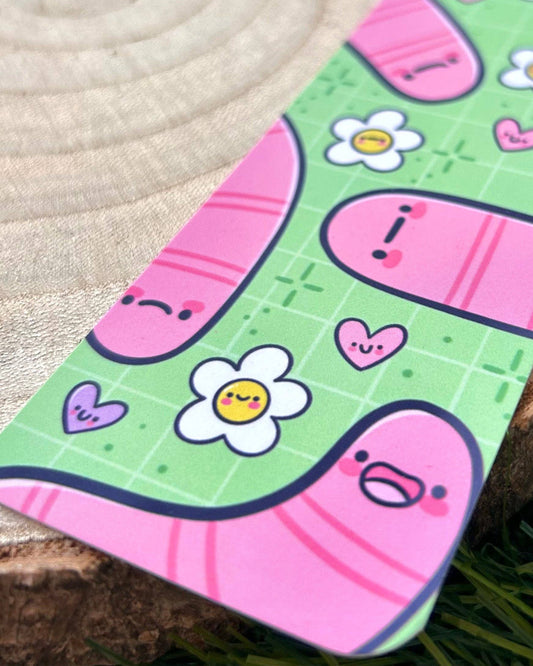 Close‑up of the Wormies Bookmark by Kelly Lou, sold by Rebel Romance. The illustrated design features pink cartoon worms with expressive faces, white daisies with smiley centres and tiny hearts on a green grid background. A playful, alternative aesthetic created by Kellylou for a small UK brand celebrating millennial style and quirky gifts.