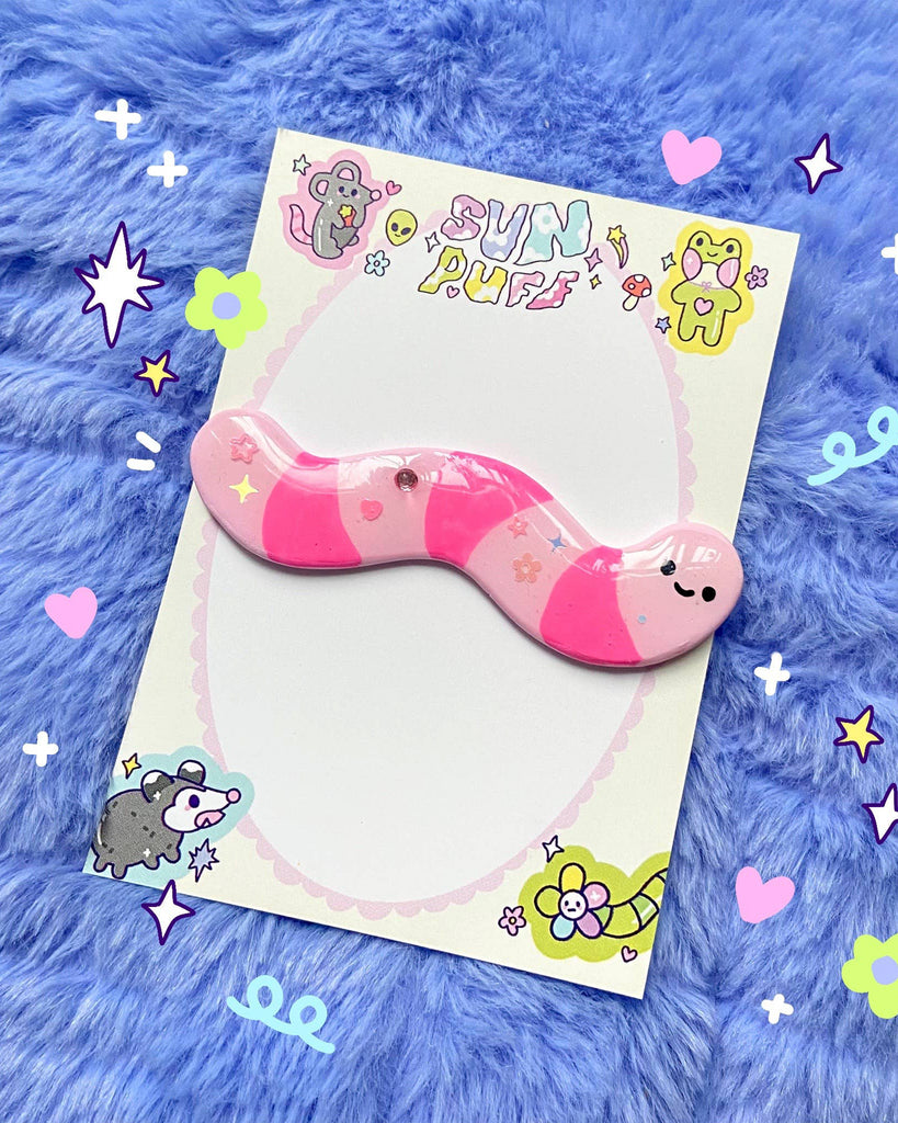 Cute handmade Wormie Hair Clip by Sun Puff Studios, available at Rebel Romance UK. This pink and pastel striped worm-shaped hair clip is crafted from polymer clay, hand-painted, sprinkled with glitter, and finished with a glossy resin coat. Attached to a silver alligator clip, it comes on a colourful backing card decorated with playful cartoon characters. A quirky accessory perfect for alternative fashion lovers and kawaii style fans.