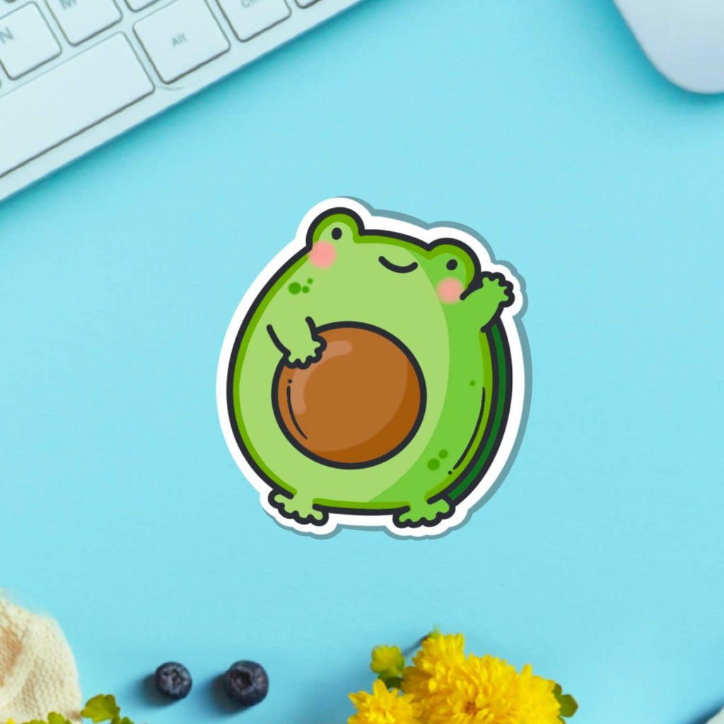 A cute vinyl sticker of a green cartoon frog with rosy cheeks, shaped like an avocado with a large brown stone in the centre. The sticker is shown on a purple background. Ideal for decorating laptops, water bottles, notebooks and more.