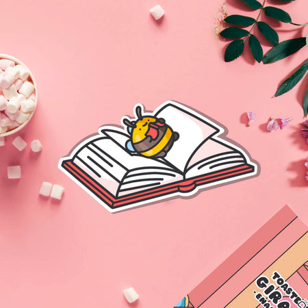 A cute vinyl sticker of a yellow and brown bumblebee lying on its back on top of an open book, tongue sticking out playfully. Displayed on a purple background with pink and blue elements. Glossy waterproof finish, ideal for book lovers and readers.