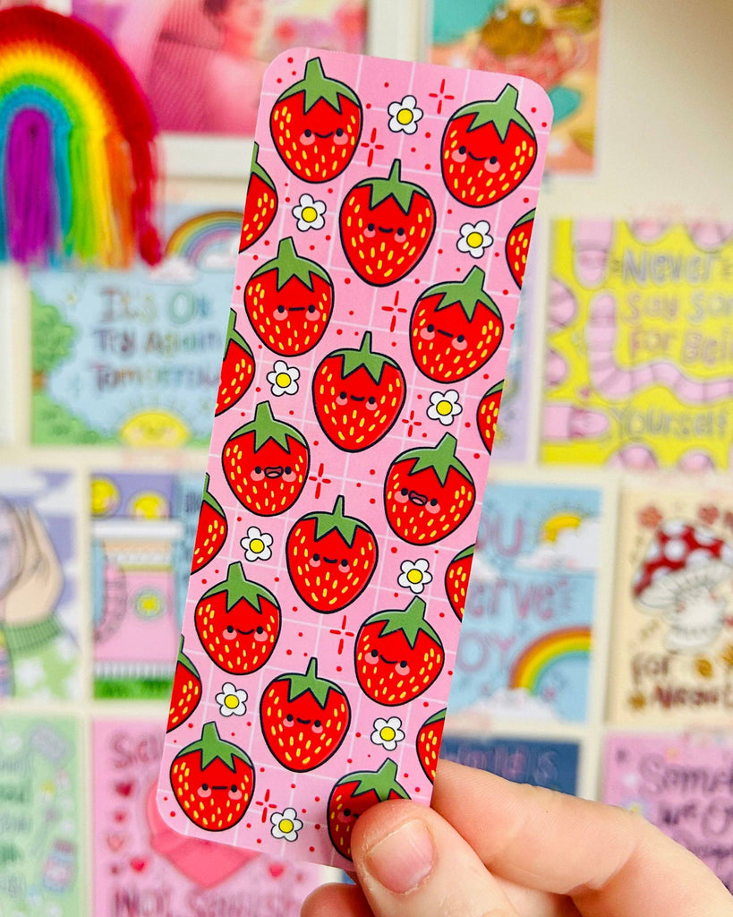 The Berry Cute Strawberries Bookmark by Kelly Lou held up in front of a colourful Rebel Romance art wall. The pink bookmark showcases smiling strawberries and daisy motifs, perfect for alternative style lovers and millennials who enjoy unique, illustrated stationery.