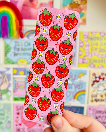 The Berry Cute Strawberries Bookmark by Kelly Lou held up in front of a colourful Rebel Romance art wall. The pink bookmark showcases smiling strawberries and daisy motifs, perfect for alternative style lovers and millennials who enjoy unique, illustrated stationery.