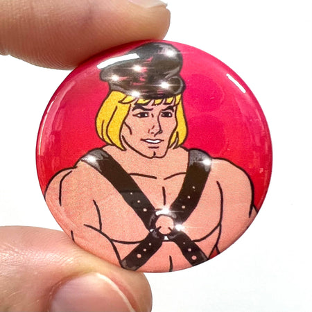 A bold and quirky button pin badge featuring a retro-inspired character resembling a blonde muscle man in a black fetish harness and cap, set against a vibrant pink background. Sold by Rebel Romance, a small UK business celebrating alternative fashion and aesthetics with unique products like cards, jewellery, and tote bags. Perfect for fans of alternative style and nostalgic designs.