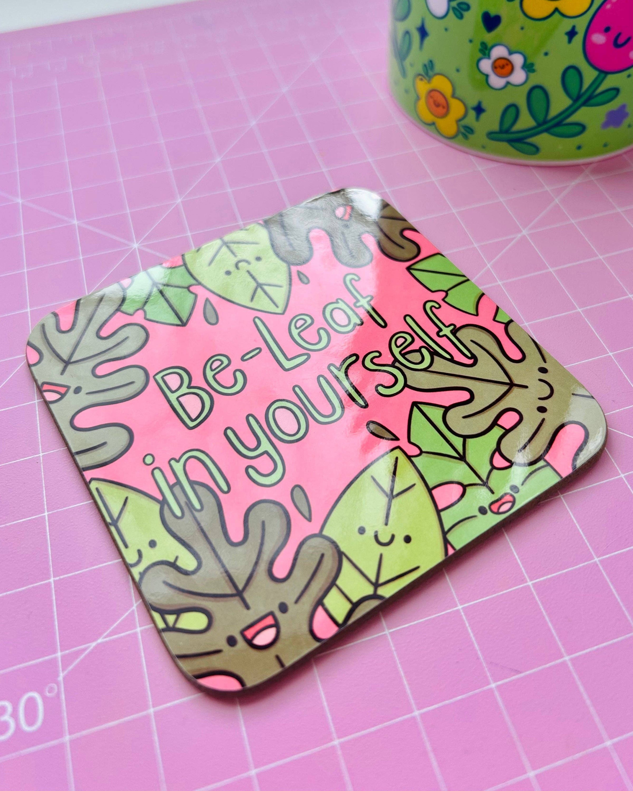 A Rebel Romance “Be‑Leaf In Yourself” drinks coaster by Kelly Lou on a pink cutting mat. The square coaster has rounded edges, a glossy finish and happy leaf illustrations, perfect as a cute, motivational gift from a small UK brand.