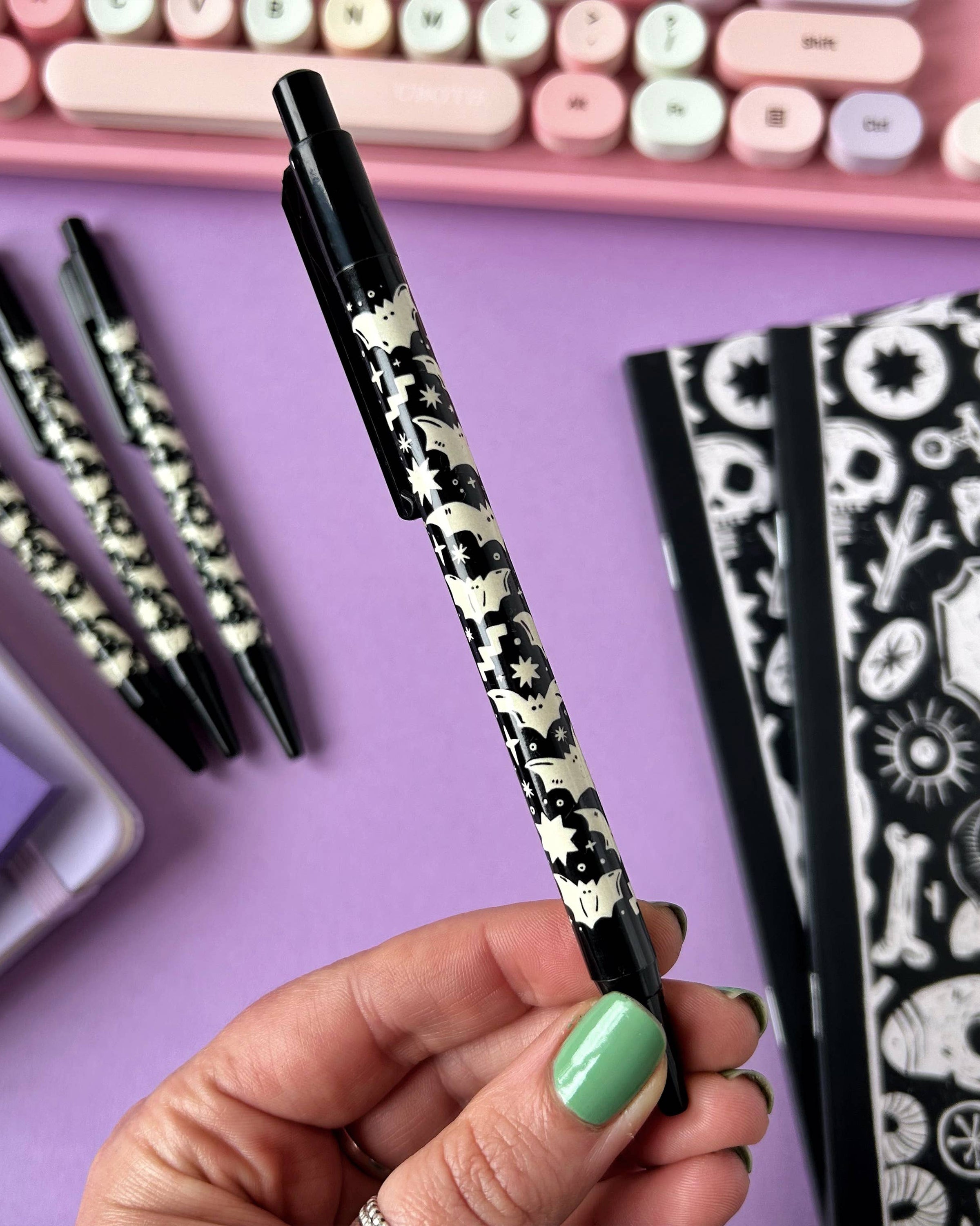 Close-up of a hand holding the Rebel Romance Bat Pattern Pen against a purple background, featuring a black and white gothic bat and star design by Stacey McEvoy Caunt, with a pastel keyboard in the background.