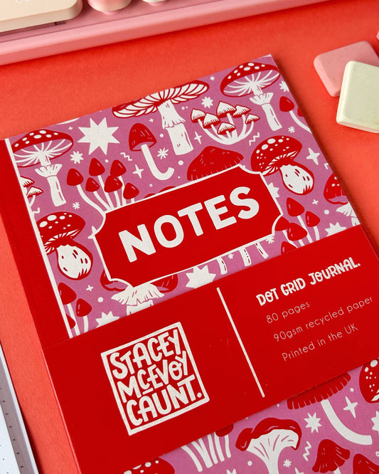 Flat lay of the pink and red mushroom notebook by Stacey McEvoy Caunt for Rebel Romance, showing the cover design with various illustrated fungi and the "NOTES" title in bold red and white. Includes visible branding and product info band.