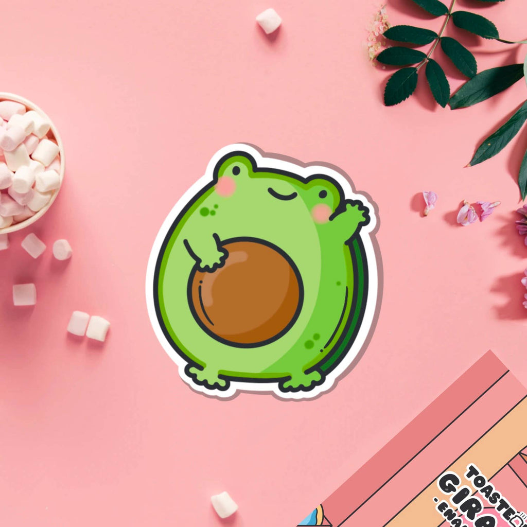 A cute vinyl sticker of a green cartoon frog with rosy cheeks, shaped like an avocado with a large brown stone in the centre. The sticker is shown on a purple background. Ideal for decorating laptops, water bottles, notebooks and more.