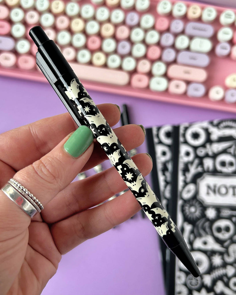 Close-up of a single Rebel Romance Bat Pattern Pen held between fingers with mint green nail polish, showing the full black and white gothic bat design and notebooks in the background.