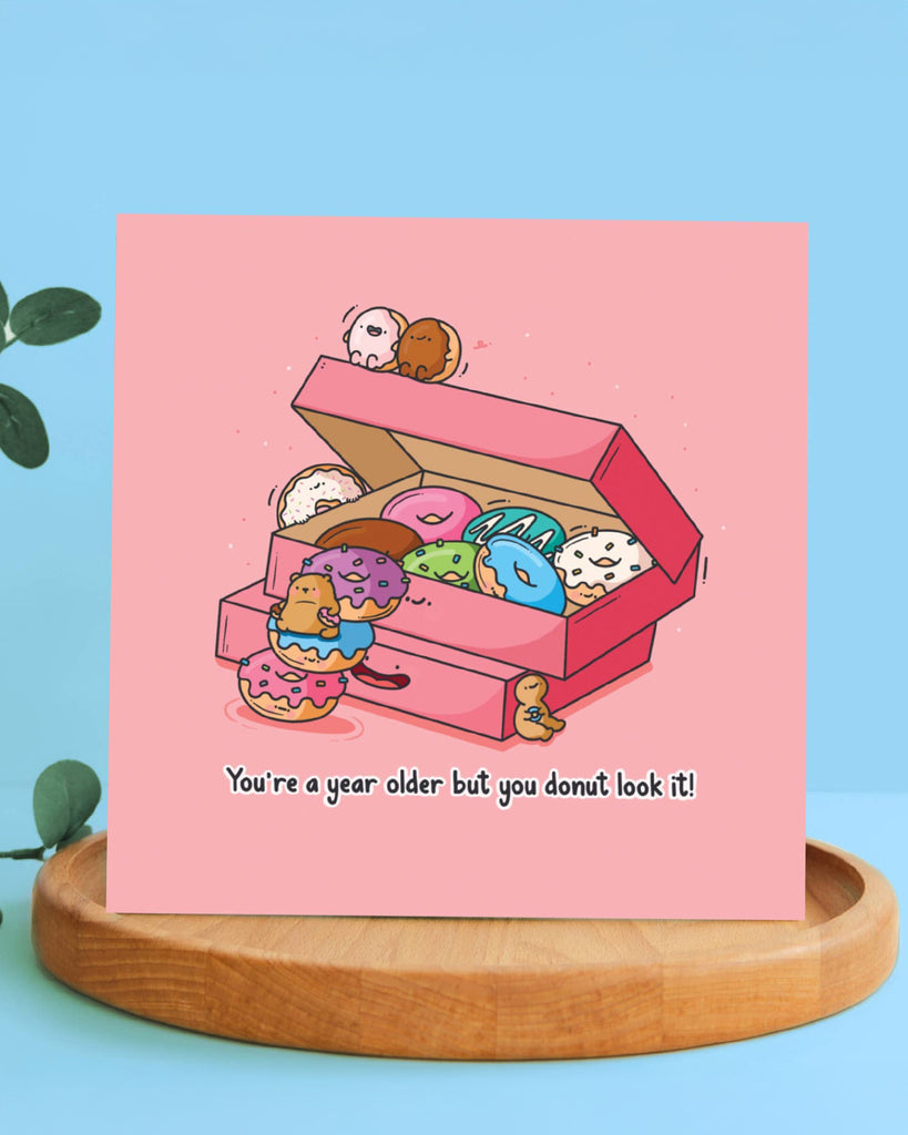 Toastedink birthday card standing on a wooden display with a light blue background, showing a pink box of kawaii-style donuts and the pun "You're a year older but you donut look it!" printed below.