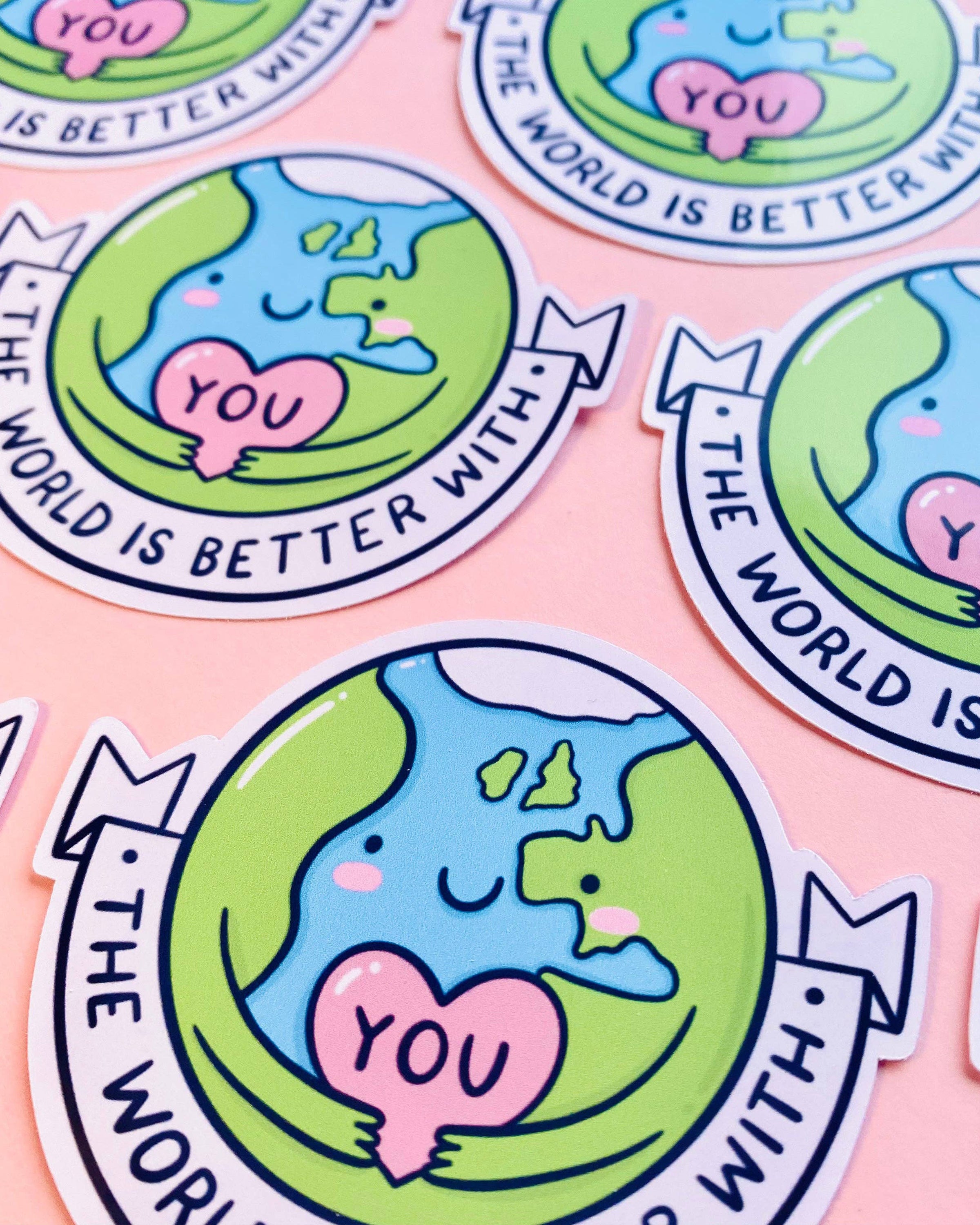 Glossy vinyl sticker from Rebel Romance featuring a cute Earth character hugging a pink heart that says "YOU", surrounded by the text “The World Is Better With You”. The design, illustrated by Kellylou, promotes self-love and positivity. Sticker is placed on a pink background. Ideal for scrapbooks, journals, and gifts.