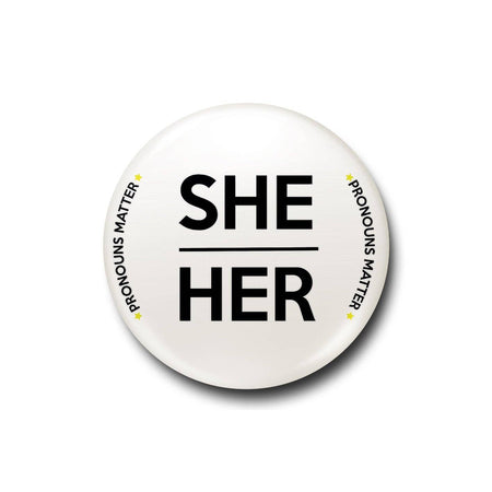 A 25mm white lapel pin featuring bold black text that reads “She/Her,” encircled by smaller curved text saying “Pronouns Matter” with yellow star accents.