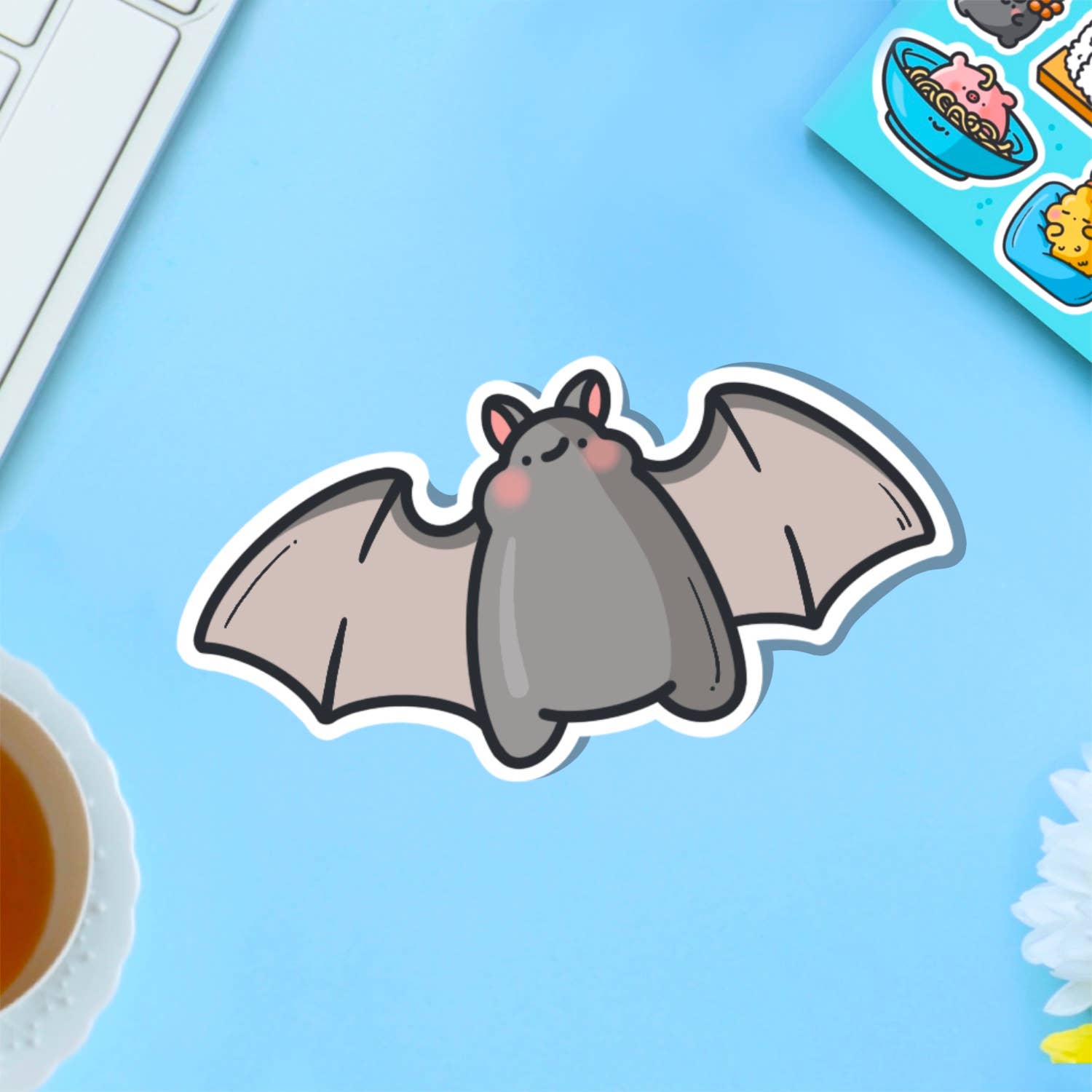 A cute vinyl sticker of a small grey cartoon bat with pink cheeks and a sweet expression, wings outstretched. Shown on a pink background with marshmallows and flowers. A waterproof, glossy sticker perfect for laptops, notebooks, or water bottles.