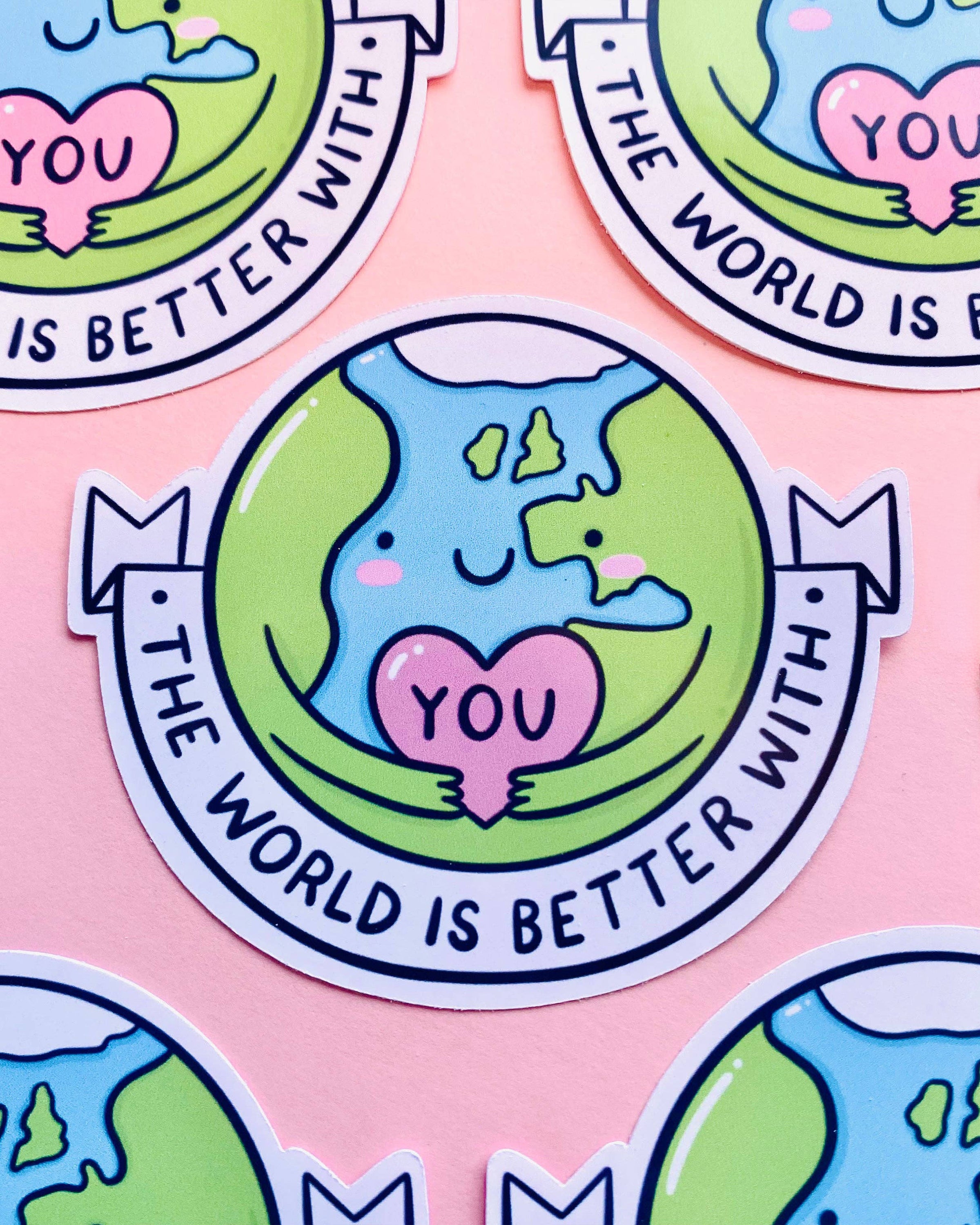 Multiple glossy Rebel Romance stickers laid out on a pink surface. Each features a cheerful cartoon Earth hugging a pink heart that reads “YOU”, with a banner that says “The World Is Better With You”. Illustrated by Kellylou, this uplifting sticker spreads positive vibes.