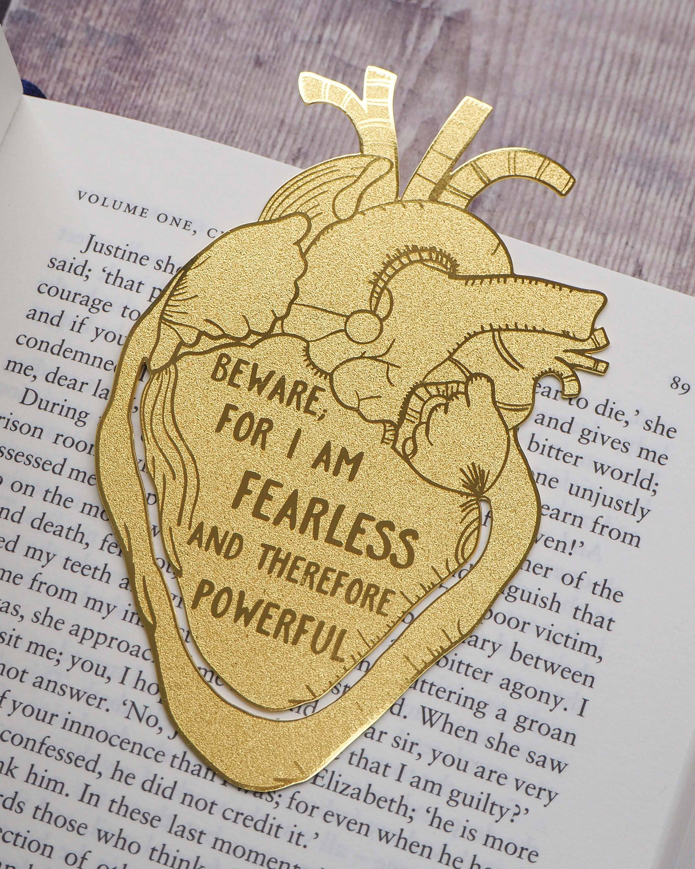 A gold brass anatomical heart-shaped bookmark engraved with the quote “Beware; for I am fearless and therefore powerful” from Mary Shelley’s Frankenstein. The bookmark is delicately etched with intricate vein details, resting on an open book to mark a page.