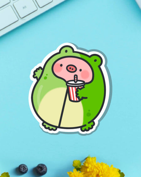 Piggy Frog vinyl sticker featuring a sweet piglet in a green frog suit, sipping a drink with a red striped straw. Pictured on a bright blue background with yellow flowers and blueberries. Sold by Rebel Romance, a UK brand for bold and whimsical gifts and stationery.