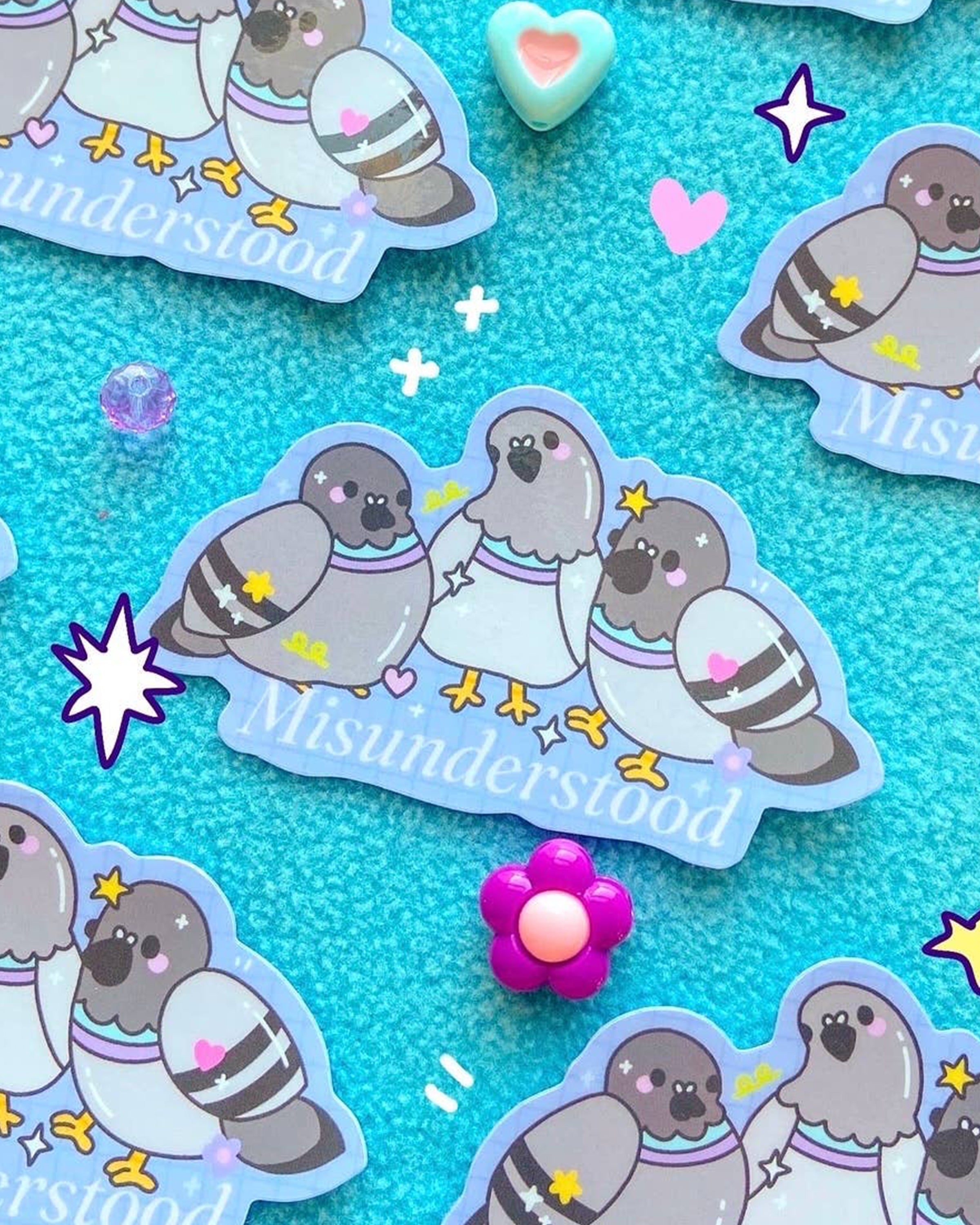 Cute vinyl sticker featuring three cartoon pigeons with pastel and 90s toy-inspired design, surrounded by sparkles and the word “Misunderstood” in white text. Illustrated by Sun Puff Studios and sold by Rebel Romance, a UK brand celebrating quirky, alternative style. Glossy finish on a bright blue background with scattered hearts, gems and playful decorations.