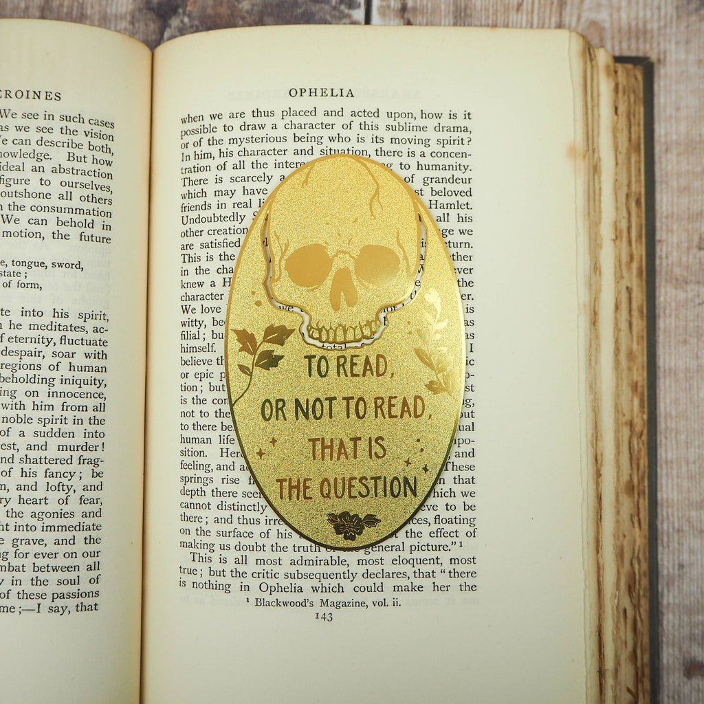 A close-up of a brass bookmark shaped like a skull, elegantly perched at the top of an open book. The intricate design of the skull is etched into the brass, giving it a striking and detailed appearance. The book’s page is titled “Ophelia,” referencing Shakespeare’s Hamlet.