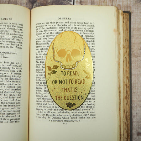 A close-up of a brass bookmark shaped like a skull, elegantly perched at the top of an open book. The intricate design of the skull is etched into the brass, giving it a striking and detailed appearance. The book’s page is titled “Ophelia,” referencing Shakespeare’s Hamlet.