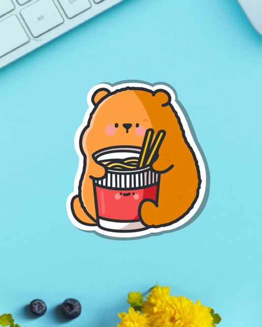 Toastedink glossy bear with ramen vinyl sticker displayed on a turquoise background next to a keyboard and flowers, showing its versatile use for planners and bottles.