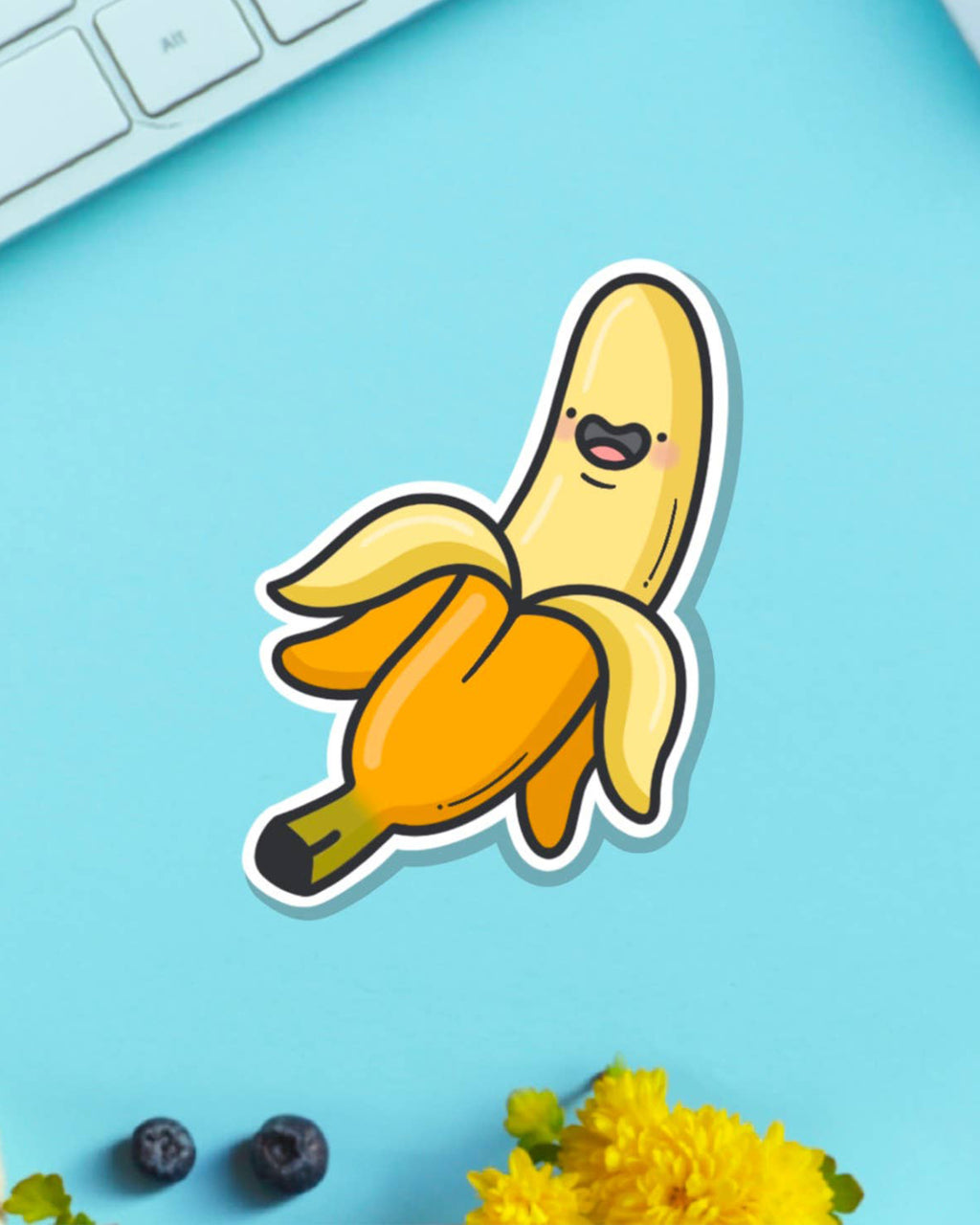 Glossy banana vinyl sticker by Toastedink, available at Rebel Romance, showing a cheerful peeled banana with a face, placed on a bright blue background with flowers and a keyboard.