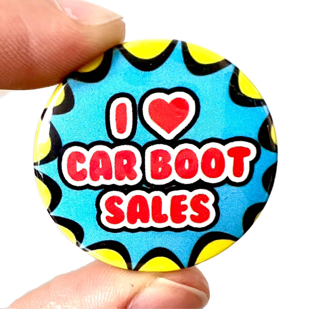 A colourful pin badge held between fingers, reading “I ❤️ Car Boot Sales” in red and white bubble text on a vibrant blue and yellow background. Designed by Bite Your Granny, available from Rebel Romance, a small UK brand known for alternative fashion, gifts, accessories, and retro-inspired homeware for millennials.