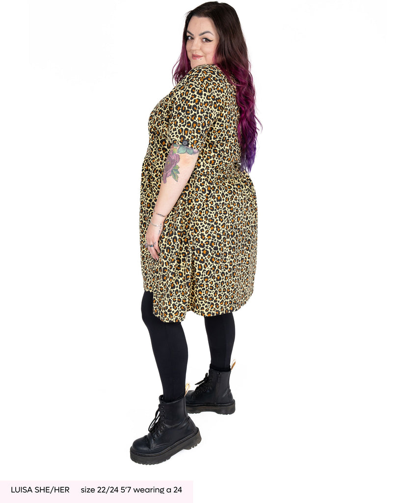 Luisa, a tattooed woman with long purple and pink hair wearing the rebel romance leopard print smock dress with black tights and black boots. She is facing left looking over her shoulder to camera smiling stood in a white studio backdrop.