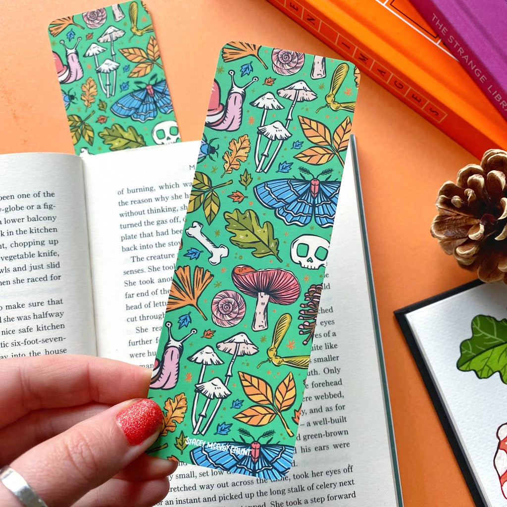 A hand with glittery red nail polish holding the green bookmark, showcasing the toad, mushrooms, and forest illustrations with an open book and autumn-themed décor in the background.