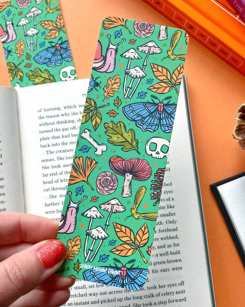 A hand with glittery red nail polish holding the green bookmark, showcasing the toad, mushrooms, and forest illustrations with an open book and autumn-themed décor in the background.
