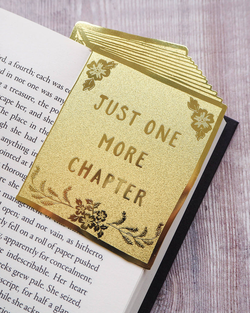 A metallic gold bookmark shaped like a book with floral detailing and the words “Just One More Chapter,” clipped onto an open novel. From Literary Emporium, available at Rebel Romance, a UK store for alternative style and unique homeware.