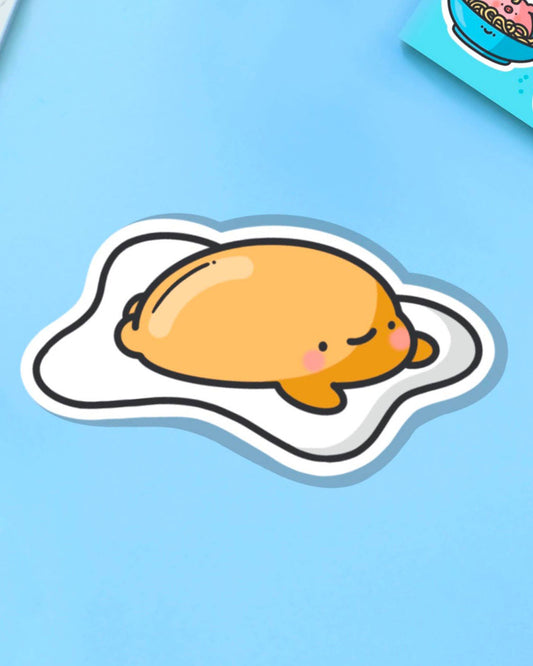 Happy fried egg sticker lounging on a glossy white base, designed by Toastedink. Set on a baby blue backdrop, this sticker is a playful addition to the Rebel Romance collection of cute, foodie-inspired accessories.