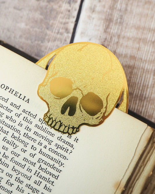 Gold brass skull bookmark shaped like a Hamlet skull clipped onto an old book page. A Literary Emporium design sold by Rebel Romance, UK creators of alternative gifts, homeware and fashion.
