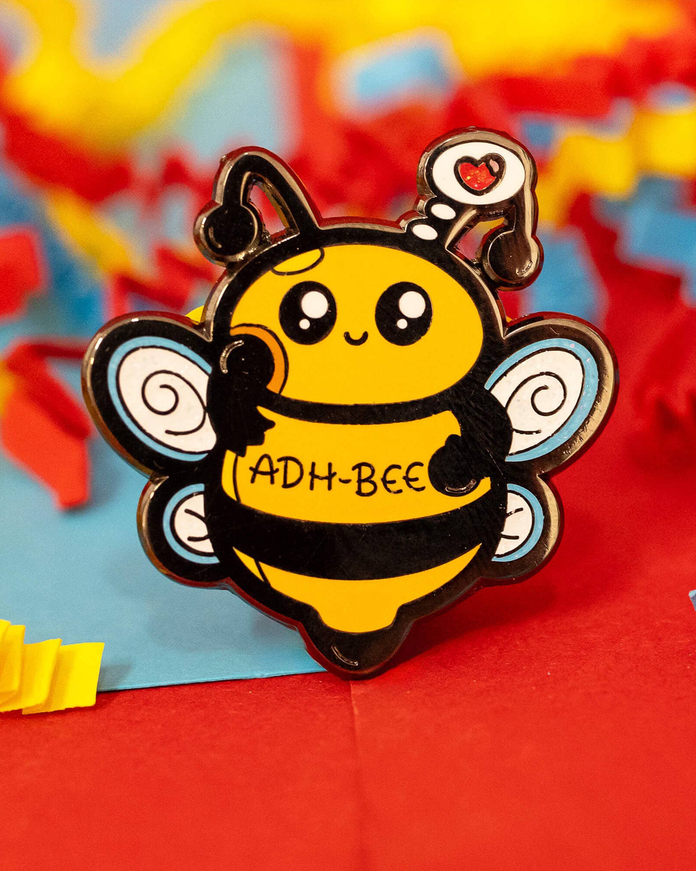 Close-up of the glittery ADH-Bee enamel pin by Innabox, showing a smiling yellow bee with big eyes, sparkly black stripes, and a heart-shaped thought bubble. Styled on a vibrant red and yellow background. Available at Rebel Romance, a UK-based alternative fashion and gift shop.