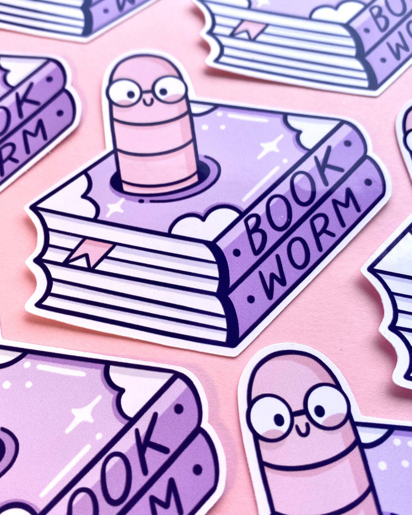 Close-up of Rebel Romance Bookworm Glossy Sticker by Kelly Lou, featuring a cute cartoon worm with glasses popping out of a purple book with the word “BOOKWORM” on the spine. Surrounded by more identical stickers on a pastel pink background. A fun, reading-themed vinyl sticker ideal for book lovers, students, and stationery fans.