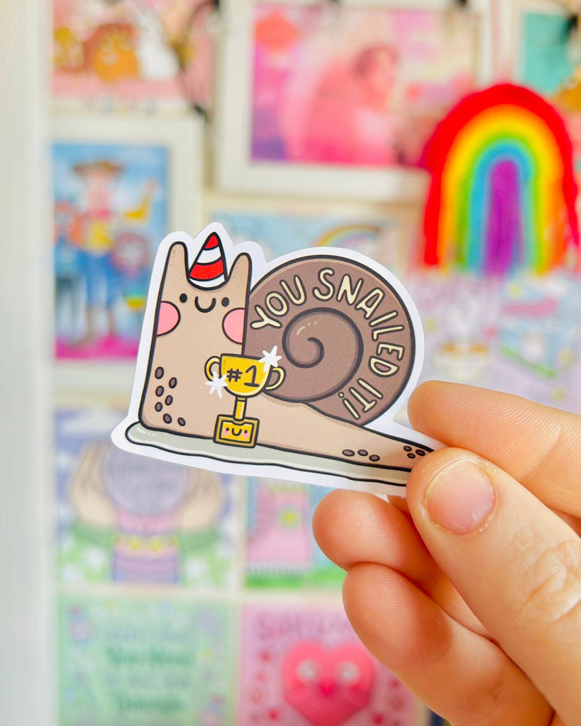 A hand holding a cute snail-shaped glossy sticker that says "You Snailed It!" with the snail wearing a party hat and holding a #1 trophy. The background is colourful and blurred with rainbows and framed artwork. Designed by Kelly Lou for Rebel Romance, a UK brand celebrating alternative, fun aesthetics with gifts, stationery, and ethical fashion.