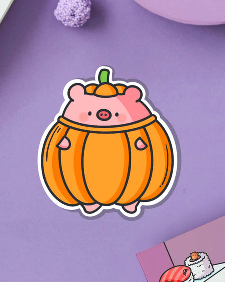 Adorable pumpkin pig vinyl sticker featuring a chubby pink pig wearing a round orange pumpkin suit with a green stem hat. Designed by Toastedink, available at Rebel Romance. Shown on a pastel purple background with cosy props. Cute waterproof sticker for seasonal or everyday fun.
