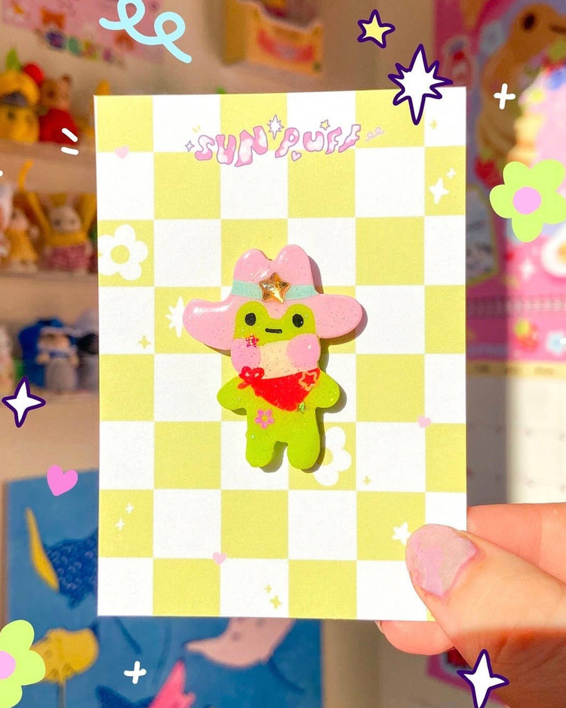 Handmade Cowboy Frog Clay Pin designed by Sun Puff Studios for Rebel Romance. The green frog character has a pink cowboy hat with glittery star, pink cheeks, red neckerchief and floral details. Mounted on a pastel yellow and white checkerboard backing card with "Sun Puff" branding. A fun kawaii accessory for alternative, playful, and millennial style aesthetics.