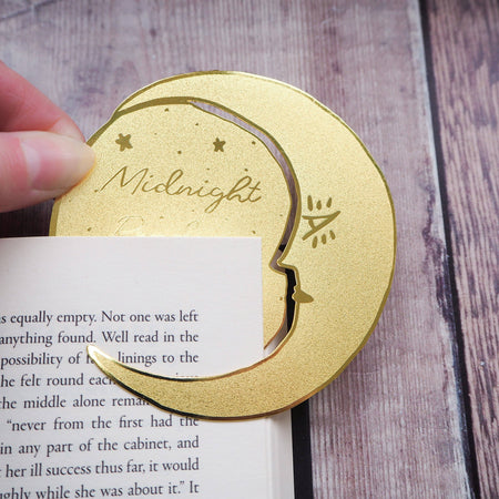 A crescent moon-shaped brass bookmark with “Midnight Readers Club” and celestial stars etched onto its surface, placed on an open book to mark a page. The bookmark adds an elegant and whimsical touch, ideal for book lovers who enjoy night-time reading.