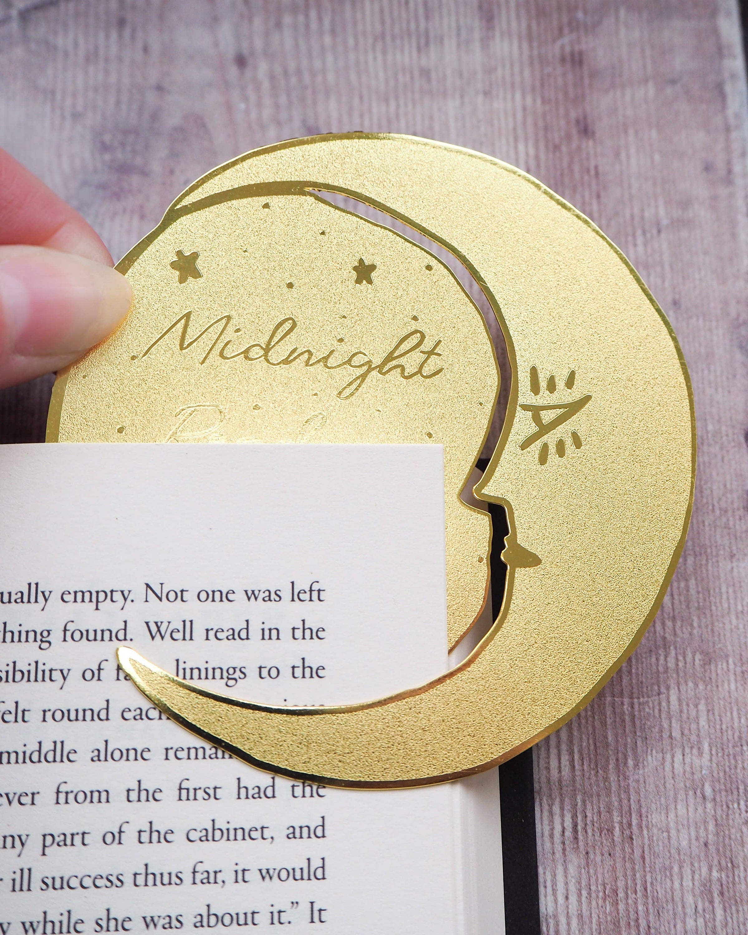 A crescent moon-shaped brass bookmark with “Midnight Readers Club” and celestial stars etched onto its surface, placed on an open book to mark a page. The bookmark adds an elegant and whimsical touch, ideal for book lovers who enjoy night-time reading.