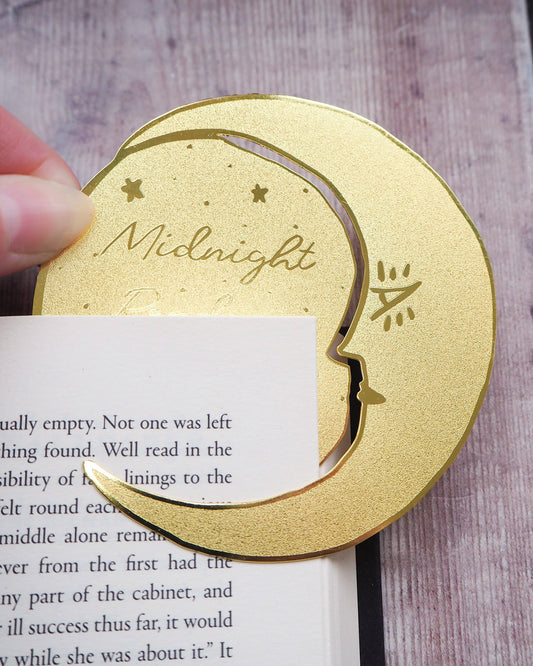 A crescent moon-shaped brass bookmark with “Midnight Readers Club” and celestial stars etched onto its surface, placed on an open book to mark a page. The bookmark adds an elegant and whimsical touch, ideal for book lovers who enjoy night-time reading.