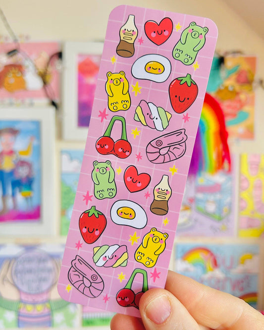 Close-up of the Pick ’n’ Mix Bookmark by Kelly Lou, sold by Rebel Romance, a small UK brand celebrating alternative style and millennial aesthetics. The pink bookmark features cute illustrated sweets with smiling faces, including gummy bears, fried eggs, cherries, marshmallows, cola bottles, hearts, strawberries and prawns, set against a grid background with stars.