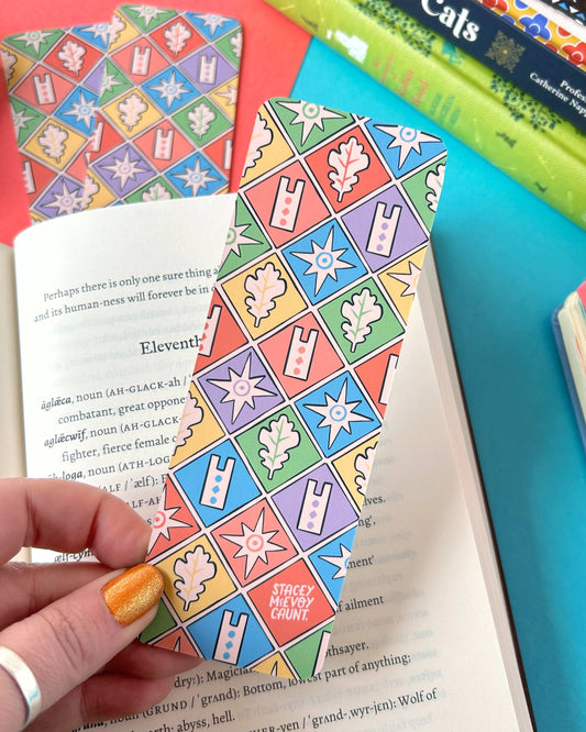 Back of the Rebel Romance Medieval Rabbits Bookmark showing a colourful medieval-inspired tile pattern with stars, oak leaves, and bold geometric shapes in red, blue, green, purple, and yellow. Held over an open book with additional bookmarks in the background.