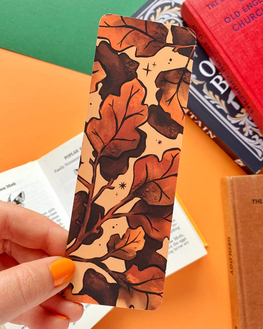 Hand holding the reverse side of the Rebel Romance Autumn Leaves Bookmark, showing a variation of the autumn leaf pattern on a beige background with subtle star accents. Surrounded by vintage books and orange paper, the setting enhances its cosy seasonal vibe.