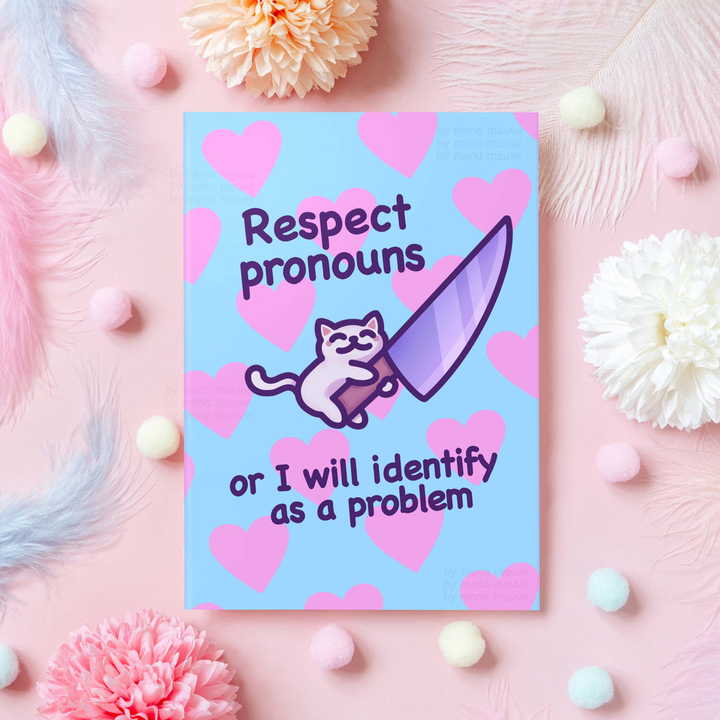 Funny LGBTQ+ greeting card with the phrase “Respect pronouns or I will identify as a problem”, featuring a cute cartoon cat holding a large pastel purple knife on a blue background with pink hearts. Displayed on a styled pink flat lay with flowers, feathers, and pom poms. Designed by Mona Mauve for Rebel Romance to support gender identity and trans visibility with humour.