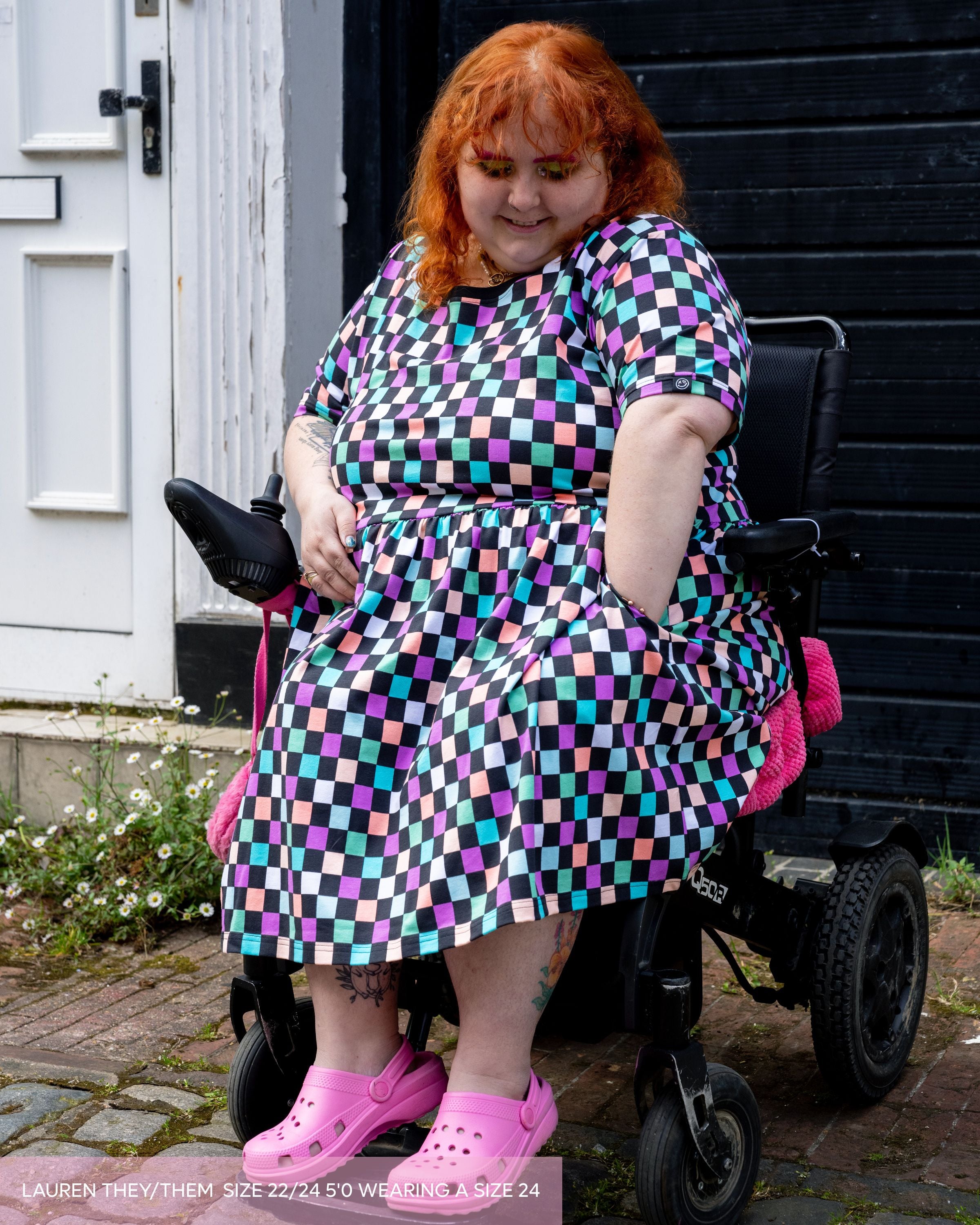 Lauren, a non binary model with curly ginger hair and brightly coloured makeup wearing the checkerboard smock dress and pink crocs sat in their mobility aid. The checkerboard print is purple, blue, orange, pink, green and pastel colours with black. 