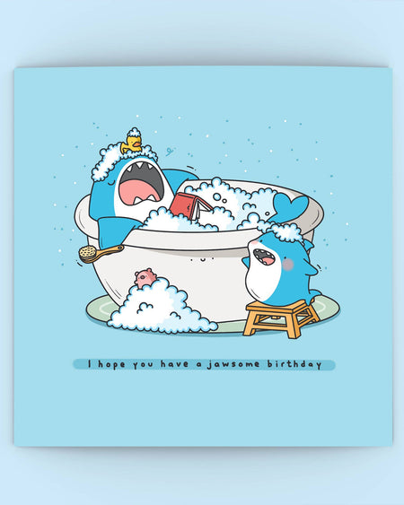 Flatlay of the Have a Jawsome Birthday card by Toastedink on a blue background. Includes a cartoon shark in a tub with bubbles and another shark beside it. A humorous, high-quality card for ocean fans, sold by Rebel Romance.