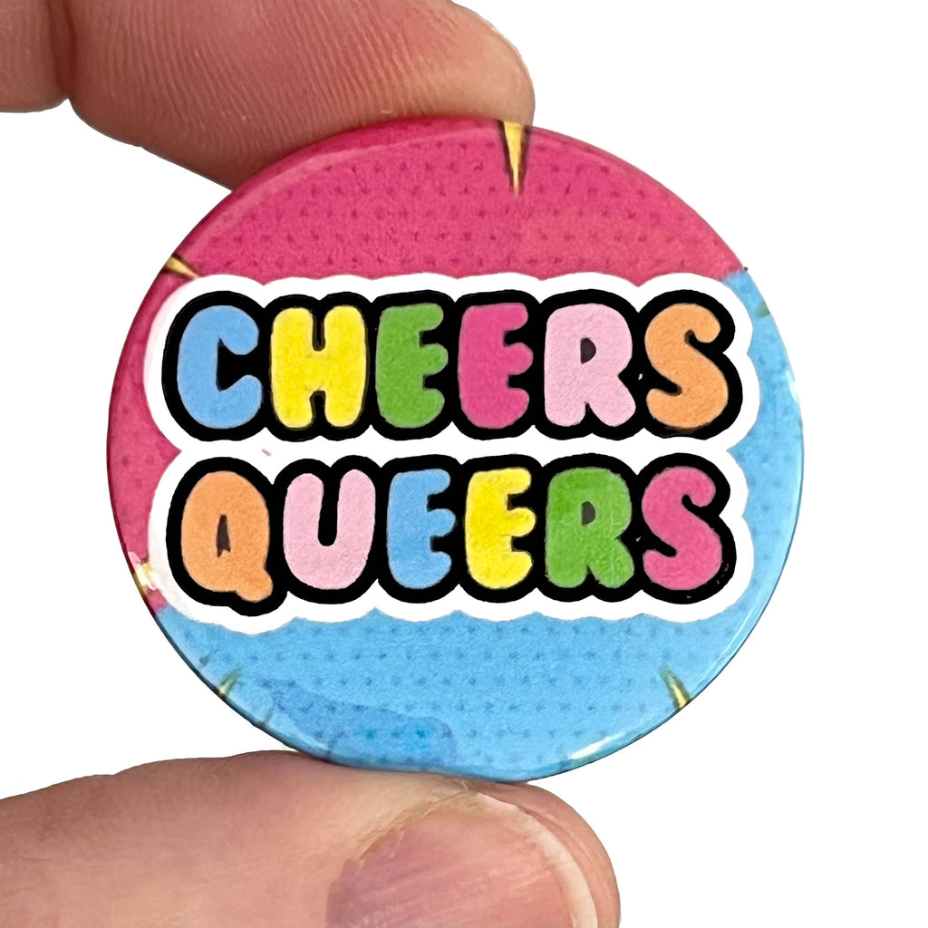 Close-up of a hand holding a Rebel Romance button pin badge with the phrase "Cheers Queers" in bold rainbow letters on a pink and blue comic-style background. Designed by Bite Your Granny, this vibrant LGBTQ+ pride badge is a fun, alternative accessory celebrating queer joy. From Rebel Romance, a small UK brand championing extraordinary style and inclusive fashion for millennials.